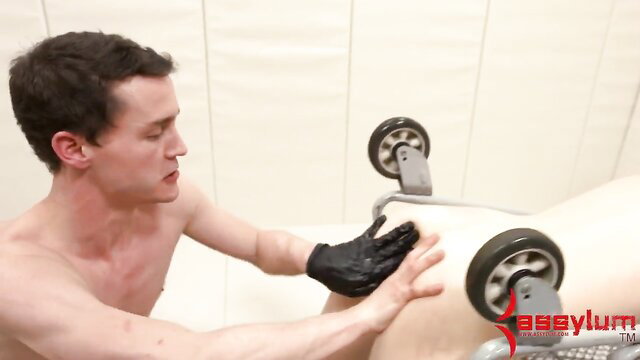 Nude man in black gloves handles wheeled bondage frame on another nude guy in gay MMF anal setup