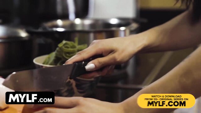 Canela Skin holding knife over bowl of green pasta in kitchen