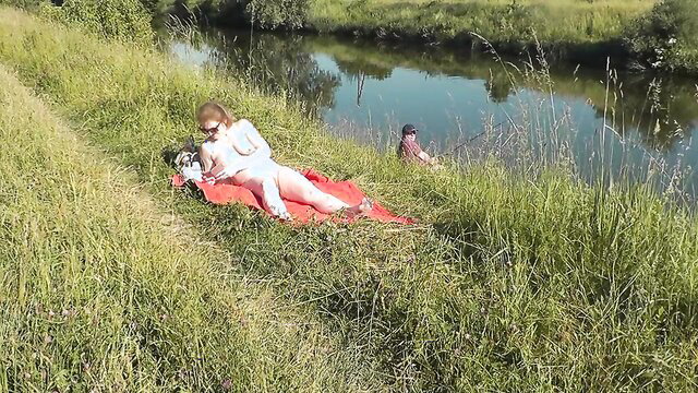 Sexy platinum blonde MILF naked sunbathing on river bank, watched by fisherman