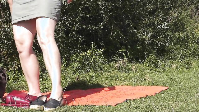 MILF in short dress and espadrilles stands on nude beach towel outdoors