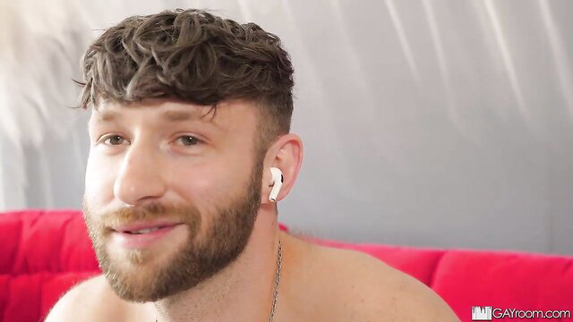 Handsome bearded white guy with curly hair smiles topless on red couch