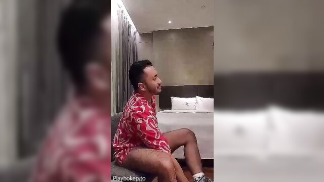 Handsome Asian guy in floral shorts sitting on hotel bed, hairy legs exposed