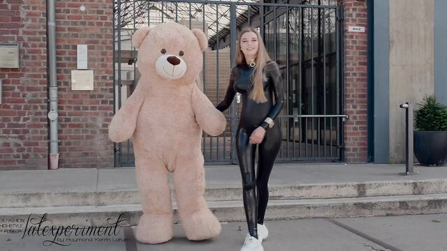 Blonde in shiny black latex catsuit hugging giant teddy bear outdoors