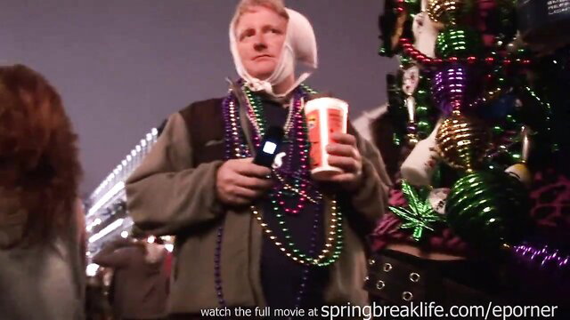 Man in headscarf with beads holds phone and drink amid Mardi Gras flashing crowd