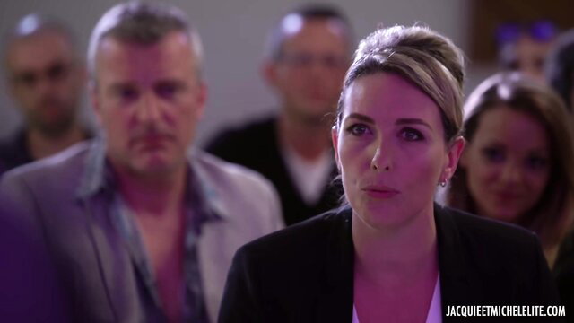 Blonde in black blazer at crowded event, tense expression, French adult scene