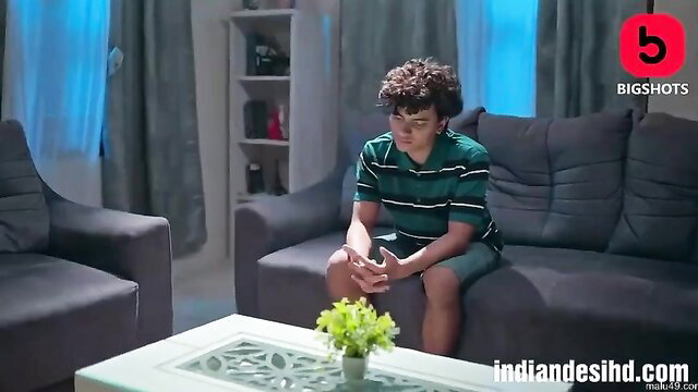 Curly-haired Indian teen boy sits pensively in shorts on couch