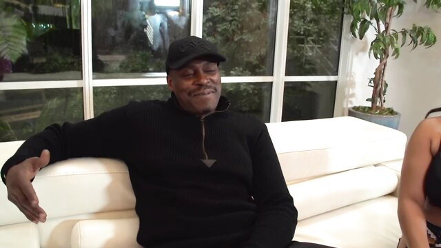 Black man grinning on couch with busty ebony woman in bra