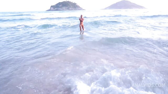 Slim Brazilian teen in bikini stands in ocean waves at beach