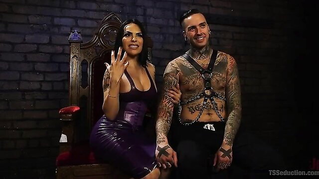 TS Foxxy in purple latex dress with tattooed slave on throne