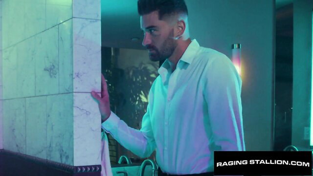 Tattooed hunk in white shirt leans against wall in Raging Stallion scene with Roman Todd