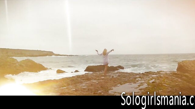 Scarlet Red arms outstretched on sunny beach rocks, solo tease