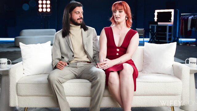 Queen Mirage in red dress with man on couch, intimate gaze