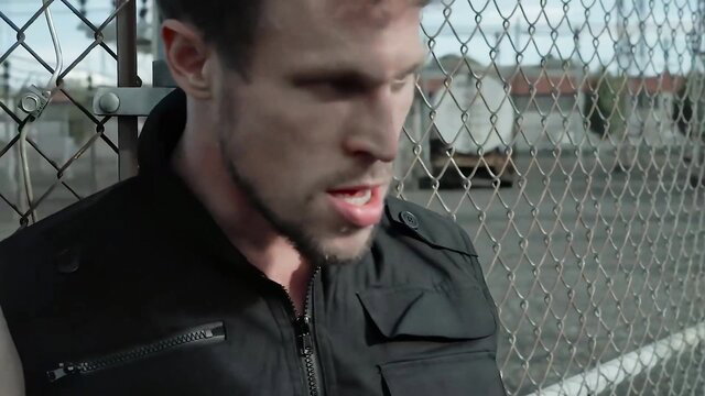 Muscular white man in tactical vest smirking behind chain link fence