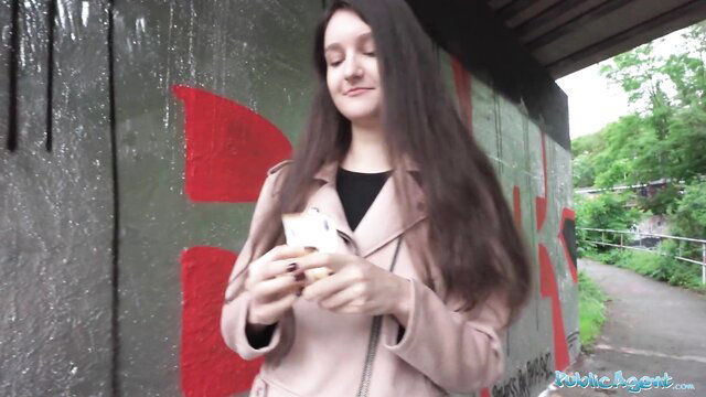 Cute 19yo European college girl in pink coat holding phone under graffiti bridge