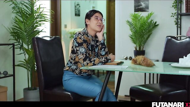 Asian T-girl in floral blouse and jeans sits pensively at table