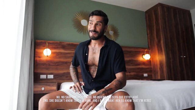 Tattooed Latino hunk in open shirt and shorts on bed, teasing bulge