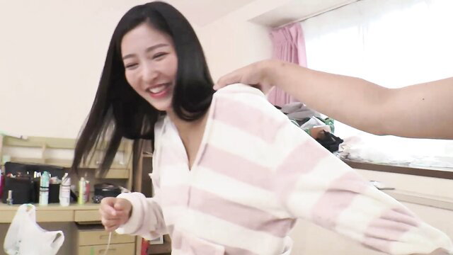 Emiri smiles playfully in pink striped robe, man groping from behind in cozy JAV scene