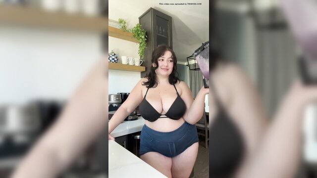 Curvy brunette in black bikini top and blue shorts posing in kitchen