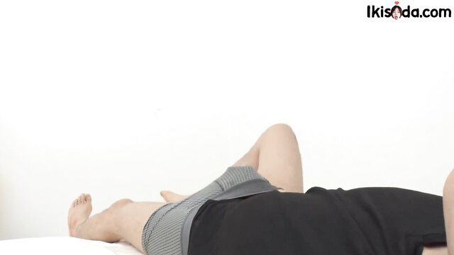 Fit young Asian guy lying naked on bed, legs spread, bulge in gray shorts