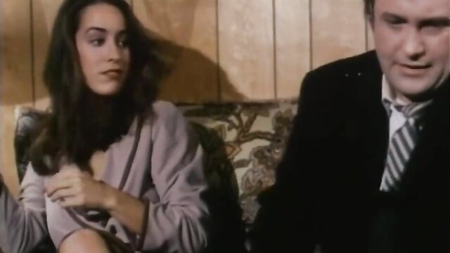 Brunette beauty in sheer robe teases beside suited man on couch