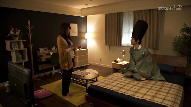 Asian woman in cardigan caring for bandaged JAV man in tall hat on bed