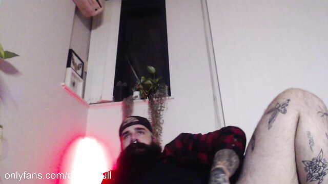 Tattooed bearded man in red plaid shirt, muscular legs spread wide