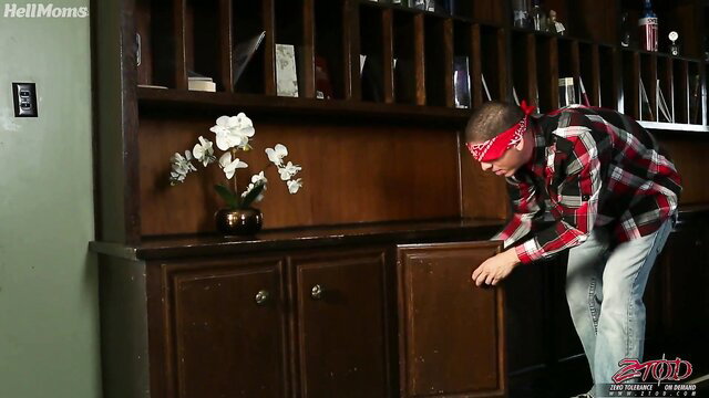 Blindfolded man in plaid shirt crouches by cabinet in MMF threesome setup