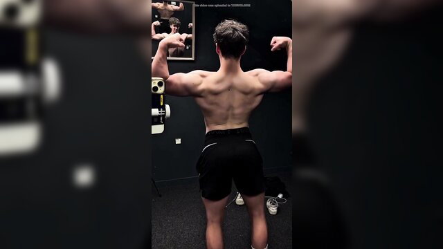 Shirtless buff white teen flexing ripped back muscles in gym shorts
