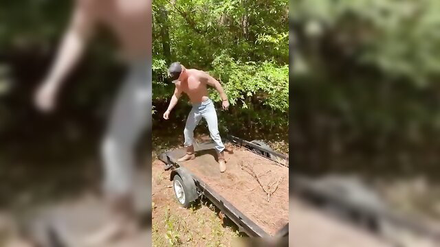 Shirtless young blonde redneck stands on trailer in woods