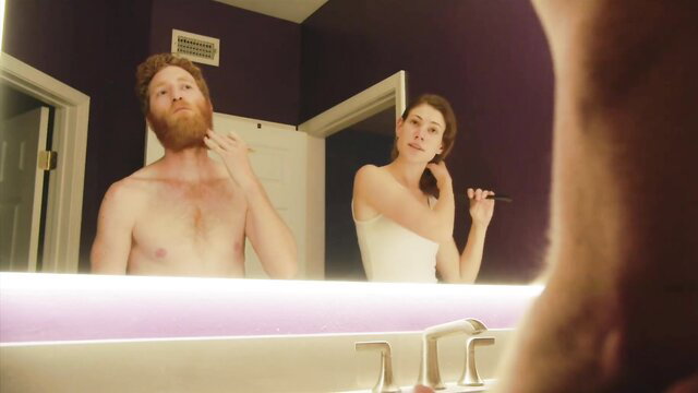 Redhead bearded hunk shirtless shaving, brunette brushing hair in mirror