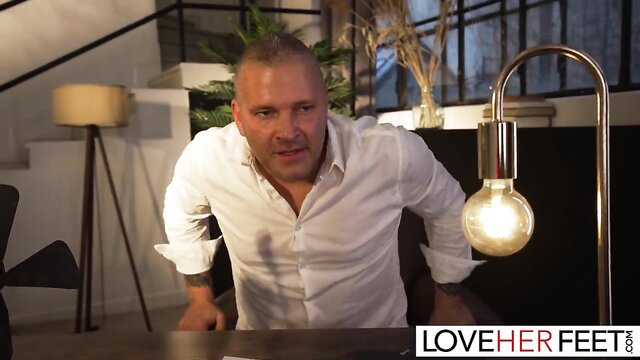 Bald muscular man in white shirt leans forward in Love Her Feet scene