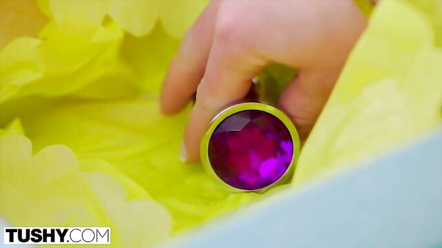 Woman holding giant purple jeweled butt plug amid yellow feathers