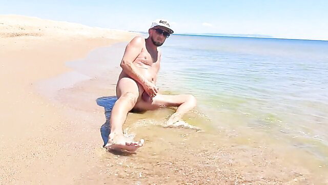 Nude bearded man sitting legs spread on sunny beach showing cock