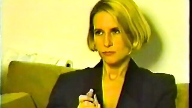 Blonde feminized maid in black suit holds object, stern gaze