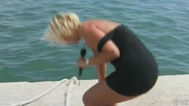 Blonde MILF in tight black dress squats by water holding mic, facefuck tease