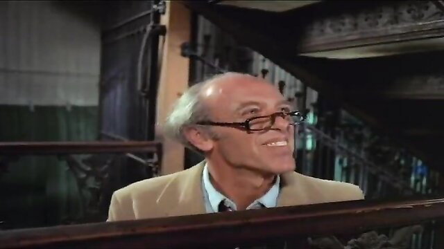 Bald older man in glasses smirks on balcony in classic Debbie Does Dallas 1978