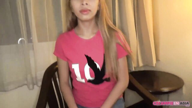 Blonde Asian in pink tee sits smiling, Thai creampie preview