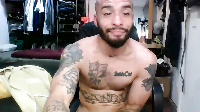 Bald muscular Latino bear with tattoos sits shirtless in gay amateur video