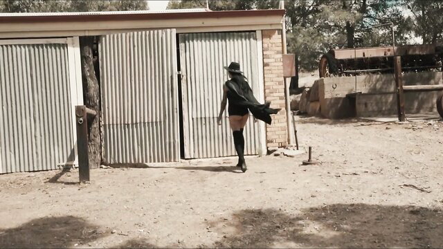 Sexy witch in cape, short skirt and stockings struts at dusty farm