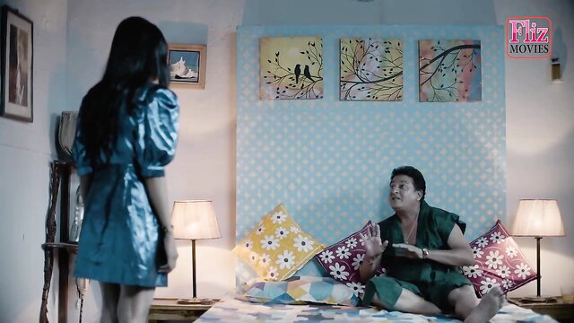 Young Indian woman in short blue dress stands before older man on bed in exotic scene