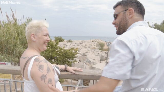 Mature blonde with big tits and tattoos chats with man outdoors by beach