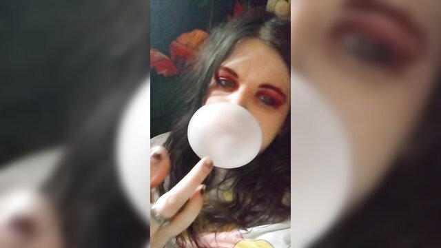 Goth girl with black hair blows huge bubble gum bubble, red eyes makeup