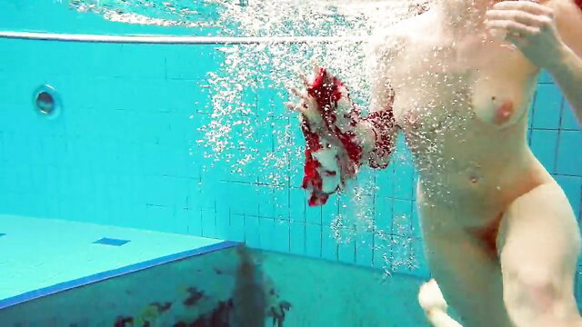 Nude redhead model swims underwater holding red attire in pool