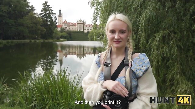 Blonde teen in pigtails by lake, off-shoulder top, camera strap, park setting