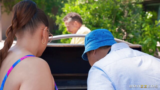 Busty Latina in bikini & man in hat bent over at outdoor BBQ