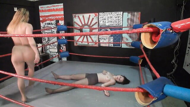 Blonde nude femdom wrestler stands over defeated Asian woman in ring