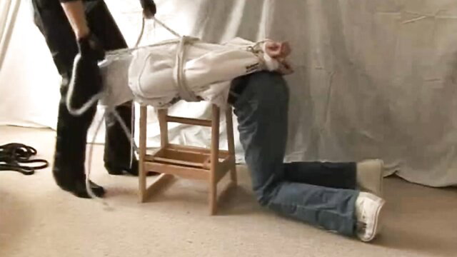 Pink boy bound in white tape and jeans, hogtied on chair in fetish bondage