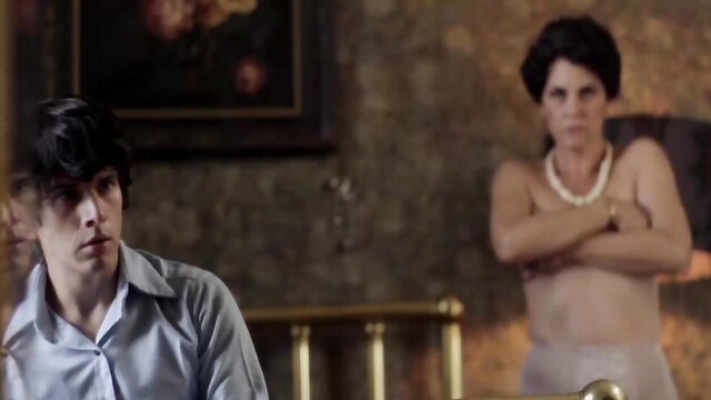 Curvy mature MILF bares huge tits, arms crossed, young man watches