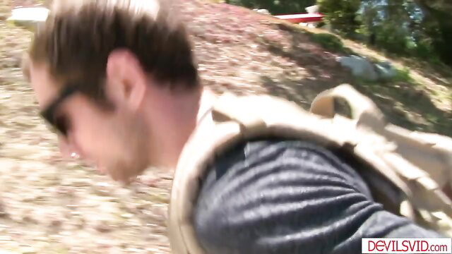 Guy in tactical vest and shades hikes near water in outdoor scene