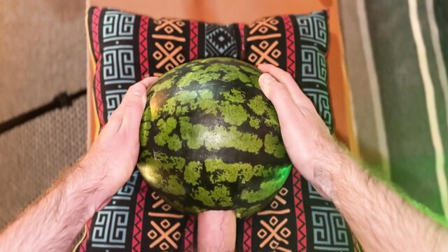 Hairy man holds green watermelon on colorful pillow in adult thumbnail
